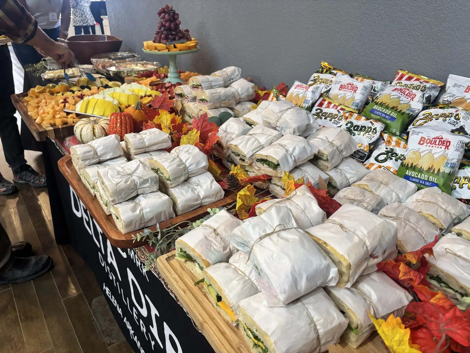 Wrapped focaccia sandwiches, chips, and a fruit tray on a buffet table